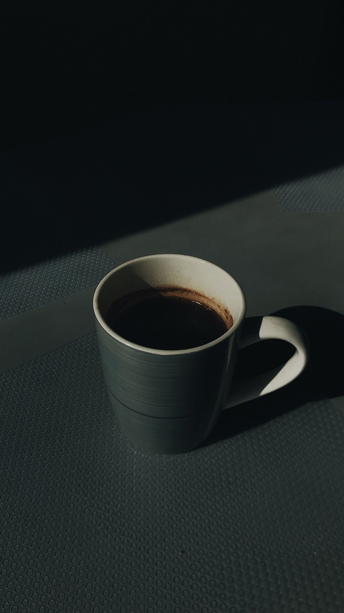 Gray ceramic mug of black coffee on a dark textured surface in a slant of light