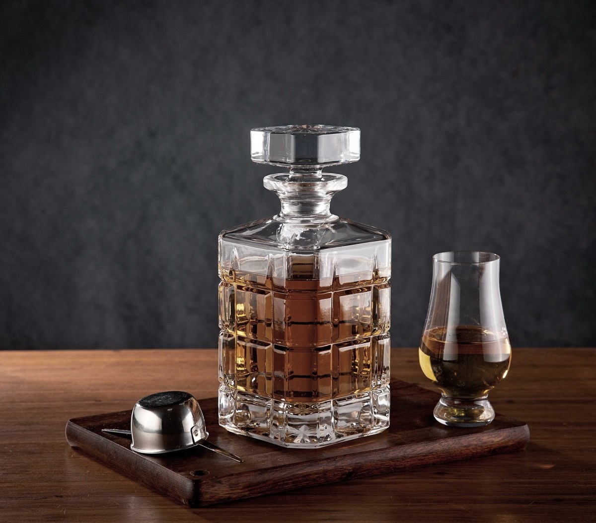Crystal whiskey decanter and tasting glass on a dark wooden tray against a gray wall