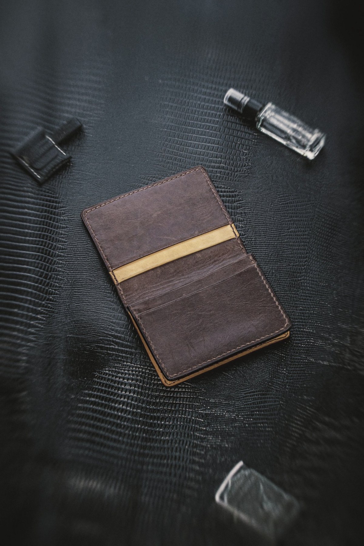 Open brown leather card holder with ochre lining on black textured leather, with small glass bottles nearby