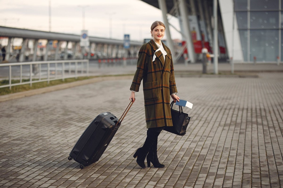 Woman in a plaid coat pulling a black suitcase outside a modern airport terminal, holding a passport